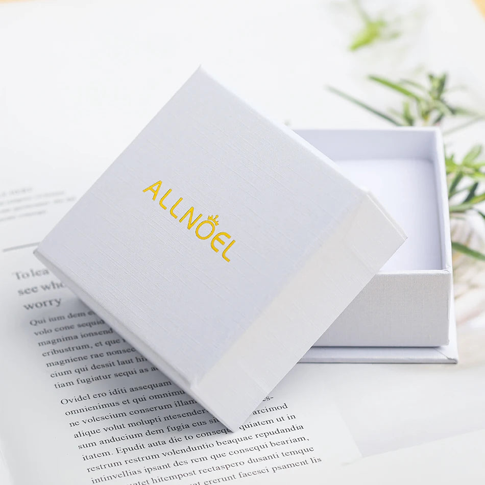 Boîte à bijoux blanche ALLNOEL pour bague en quartz rose, posée sur un livre ouvert avec décor floral.